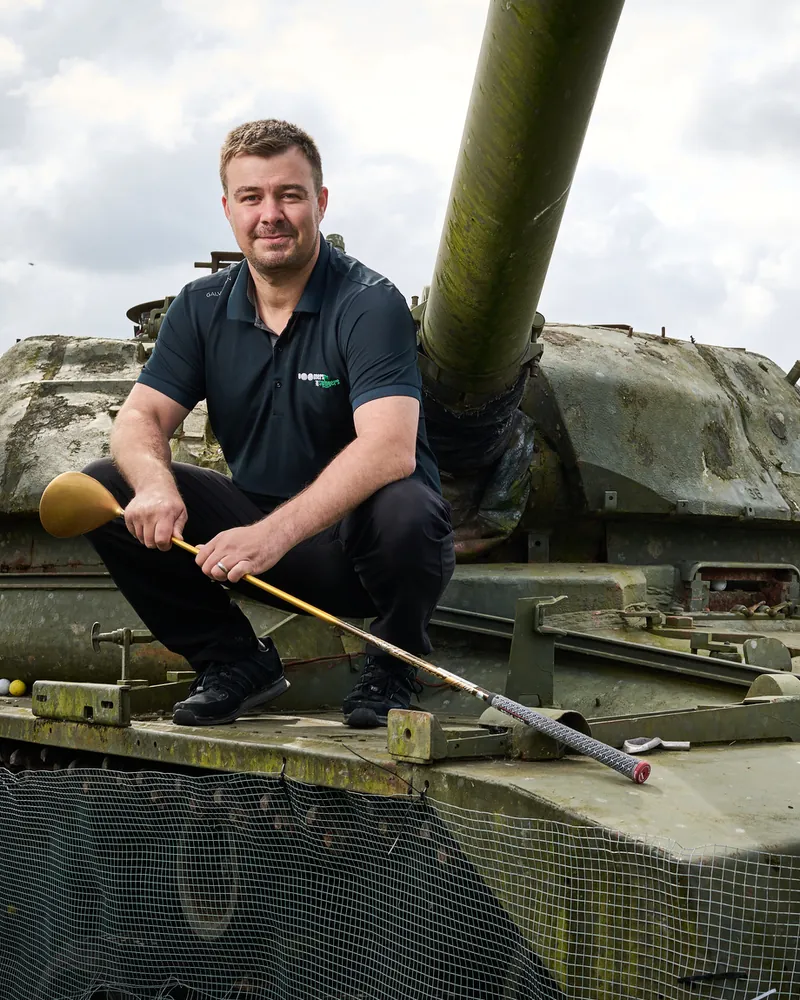 Nick Solski, smiling, crouched on the old tank at Boomers and Swingers driving range with a driver in his hands